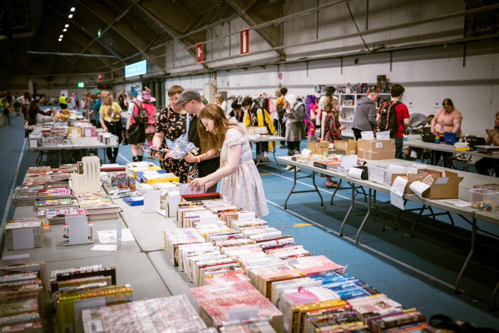 Kirpputoripöytä Kotae Expossa. Etualalla mangapokkareita, taustalla nainen katselemassa pöydän tarjontaa.